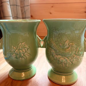 May include: Two light green ceramic vases with a glossy finish. Each vase has a bulbous body, a flared rim, and a pedestal base. One vase features a floral relief design, while the other has a crackle glaze and a bird relief.