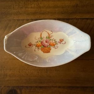 May include: A small, oval-shaped dish with a light purple glaze. The dish has a floral design with a basket of flowers in the center.