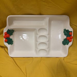 May include: White ceramic serving tray with four compartments, including a small oval section with five indentations. The tray has green holly leaf accents with red berries on each side.
