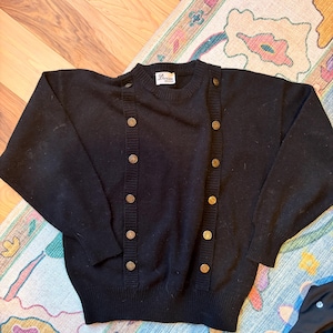 May include: Black sweater with gold-colored buttons. The sweater has a crew neck and long sleeves. The buttons are arranged in vertical rows on the front. The label on the sweater reads "Durian".
