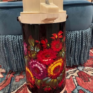 May include: Vintage insulated beverage container with a cream-colored top and a black body. The body is decorated with a vibrant floral pattern featuring red, yellow, and purple flowers with green leaves. The container has a handle and a push-button dispenser.