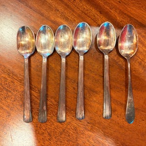 May include: A set of six silver-toned metal spoons arranged in a row. Each spoon features an oval-shaped bowl and a straight handle with a series of vertical, parallel lines etched along the length of the stem, providing a textured appearance.