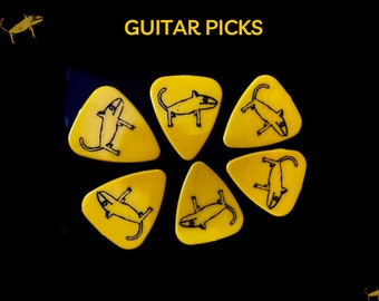 May include: Six yellow guitar picks with black line drawings of a cartoon lizard on each pick. The picks are arranged in a circle on a black background. The text "GUITAR PICKS" is written in yellow letters at the top of the image.