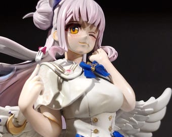 May include: A detailed figurine of a female anime character with light purple hair and yellow eyes. She wears a white dress with blue bows and has white angel wings. The figure is winking and smiling.
