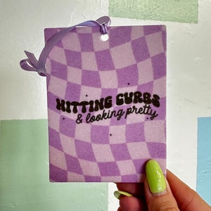 May include: Purple and white checkered car air freshener with the text "Hitting curves & looking pretty".