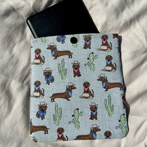 May include: A light blue fabric case with a pattern of brown dachshunds wearing cowboy hats and bandanas. The dachshunds are surrounded by cacti and small white dots. A black e-reader is partially visible inside the case.