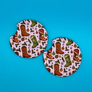 May include: Two round car coasters with a light pink background. The coasters feature a repeating pattern of brown, green, and red cowboy boots, Christmas ornaments, and holly leaves. The coasters are designed to fit in a car's cup holder.