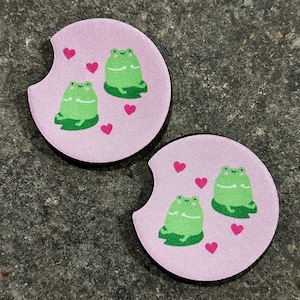 May include: Two pink round coasters with a white border. Each coaster features a green frog with a heart design. The frogs are sitting on a green leaf. The coasters have a cut-out section for a cup.