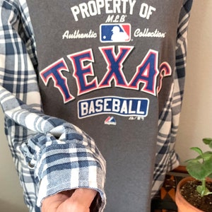 Puede incluir: Camiseta gris con un logotipo de "Texas Baseball" azul y rojo. La camiseta tiene una camisa de franela a cuadros blanco y azul superpuesta. El logotipo dice "Property of MLB Authentic Collection".
