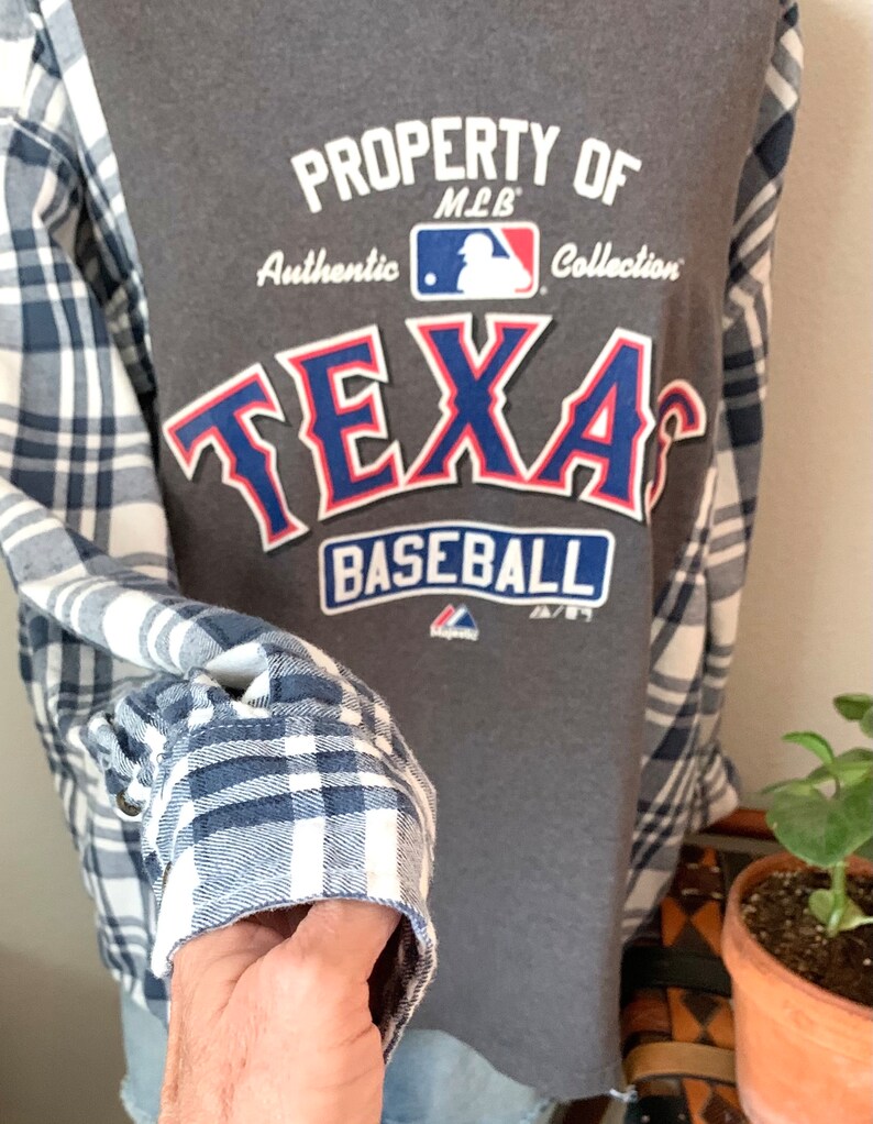 Puede incluir: Camiseta gris con un logotipo de "Texas Baseball" azul y rojo. La camiseta tiene una camisa de franela a cuadros blanco y azul superpuesta. El logotipo dice "Property of MLB Authentic Collection".