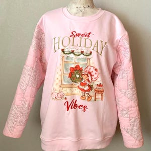 May include: Pink sweatshirt with a quilted design on the sleeves. The front features a graphic of Strawberry Shortcake holding a Christmas wreath with the text "Sweet Holiday Vibes".