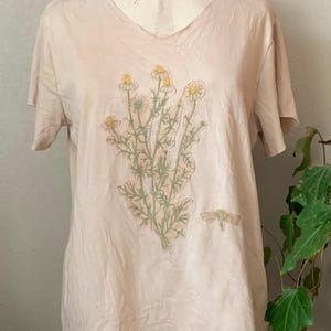 Puede incluir: Una camiseta rosa claro con un dise&ntilde;o floral que presenta un grupo de flores amarillas y hojas verdes. El dise&ntilde;o est&aacute; centrado en la parte delantera de la camiseta.