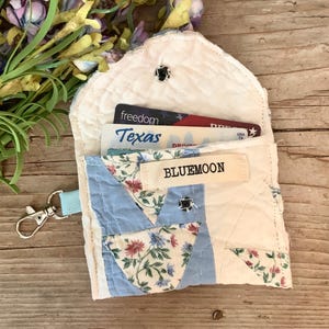 May include: A handmade, quilted wallet with a light blue strap and a silver-colored clasp. The wallet is off-white with blue and floral patterned fabric accents. The word "BLUEMOON" is printed on a fabric tag. Inside are cards.