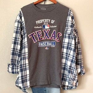 Puede incluir: Camiseta gris con un logotipo de "Texas Baseball" azul y rojo. La camiseta est&aacute; superpuesta a una camisa de franela a cuadros azules y blancos.