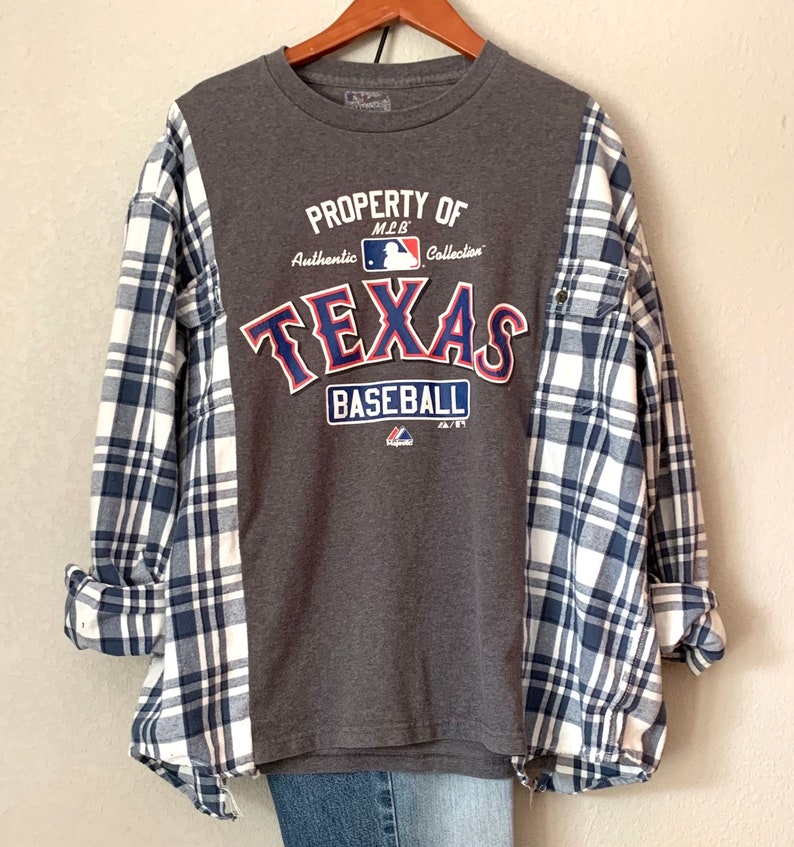 Puede incluir: Camiseta gris con un logotipo de "Texas Baseball" azul y rojo. La camiseta est&aacute; superpuesta a una camisa de franela a cuadros azules y blancos.