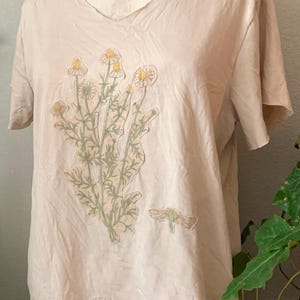 Puede incluir: Una camiseta rosa p&aacute;lida con un dise&ntilde;o floral que presenta un grupo de margaritas blancas con centros amarillos y hojas verdes. Tambi&eacute;n se puede ver un peque&ntilde;o insecto verde en la camiseta.