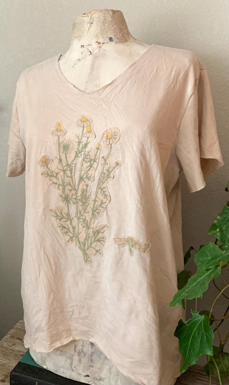 Puede incluir: Una camiseta rosa p&aacute;lida con un dise&ntilde;o floral que presenta un grupo de margaritas blancas con centros amarillos y hojas verdes. Tambi&eacute;n se puede ver un peque&ntilde;o insecto verde en la camiseta.