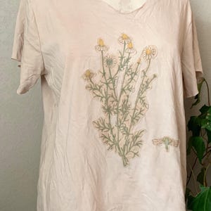 Puede incluir: Una camiseta rosa claro con un dise&ntilde;o floral que presenta un grupo de flores blancas con centros amarillos y hojas verdes. El dise&ntilde;o est&aacute; centrado en la parte delantera de la camiseta.