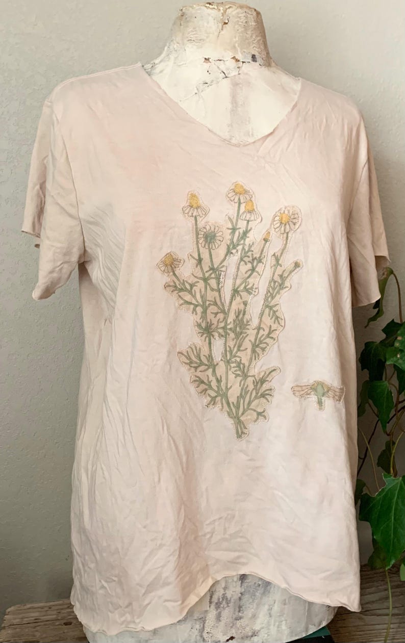 Puede incluir: Una camiseta rosa claro con un dise&ntilde;o floral que presenta un grupo de flores blancas con centros amarillos y hojas verdes. El dise&ntilde;o est&aacute; centrado en la parte delantera de la camiseta.