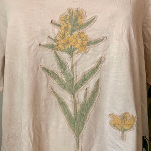 Puede incluir: Una camiseta beige claro con un dise&ntilde;o de flor amarilla. La flor est&aacute; hecha de tela y tiene hojas verdes.