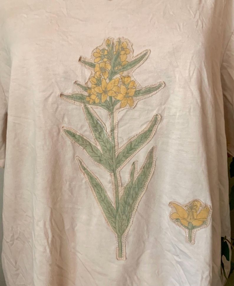 Puede incluir: Una camiseta beige claro con un dise&ntilde;o de flor amarilla. La flor est&aacute; hecha de tela y tiene hojas verdes.