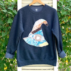 May include: A dark blue sweatshirt with a patchwork witch hat design. The hat is made of various colorful fabrics and has a frayed edge.