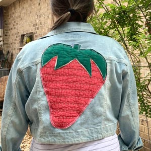 May include: A light blue denim jacket with a red and green quilted strawberry patch sewn on the back.
