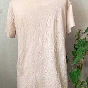 Puede incluir: Una camiseta de color beige claro con un escote ligeramente desgastado. La camiseta est&aacute; hecha de una tela suave y texturizada.