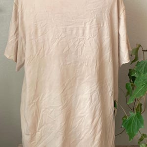 Puede incluir: Una camiseta lisa de color blanco roto, de manga corta, con una textura ligeramente arrugada. La camiseta est&aacute; hecha de una tela suave y ligera.