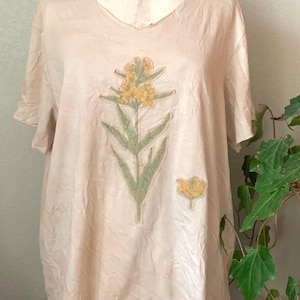 Puede incluir: Una camiseta beige claro con un estampado floral de flores amarillas y hojas verdes. La camiseta tiene cuello en V y mangas cortas.
