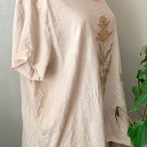Puede incluir: Una camiseta beige p&aacute;lida con un dise&ntilde;o floral impreso en la parte delantera. El dise&ntilde;o presenta un solo tallo con m&uacute;ltiples flores amarillas y hojas verdes.