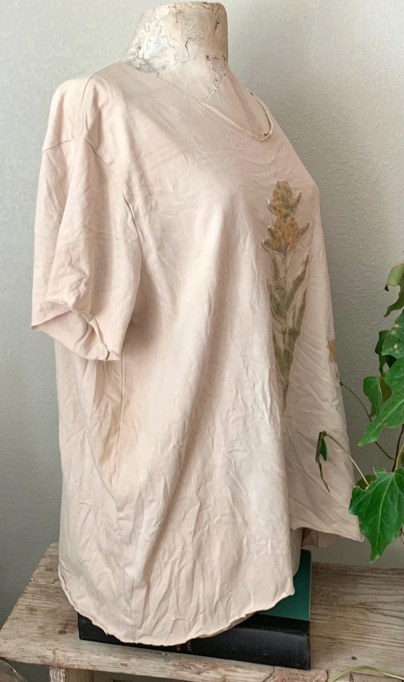 Puede incluir: Una camiseta beige p&aacute;lida con un dise&ntilde;o floral impreso en la parte delantera. El dise&ntilde;o presenta un solo tallo con m&uacute;ltiples flores amarillas y hojas verdes.