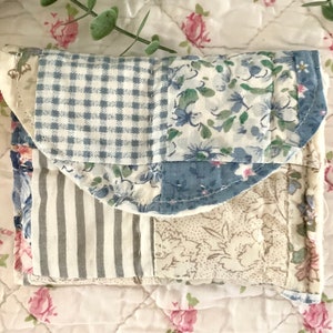 May include: A small, quilted fabric pouch with a patchwork design. The pouch is made of different fabrics in shades of blue, white, and gray. The fabric is patterned with flowers, stripes, and gingham.