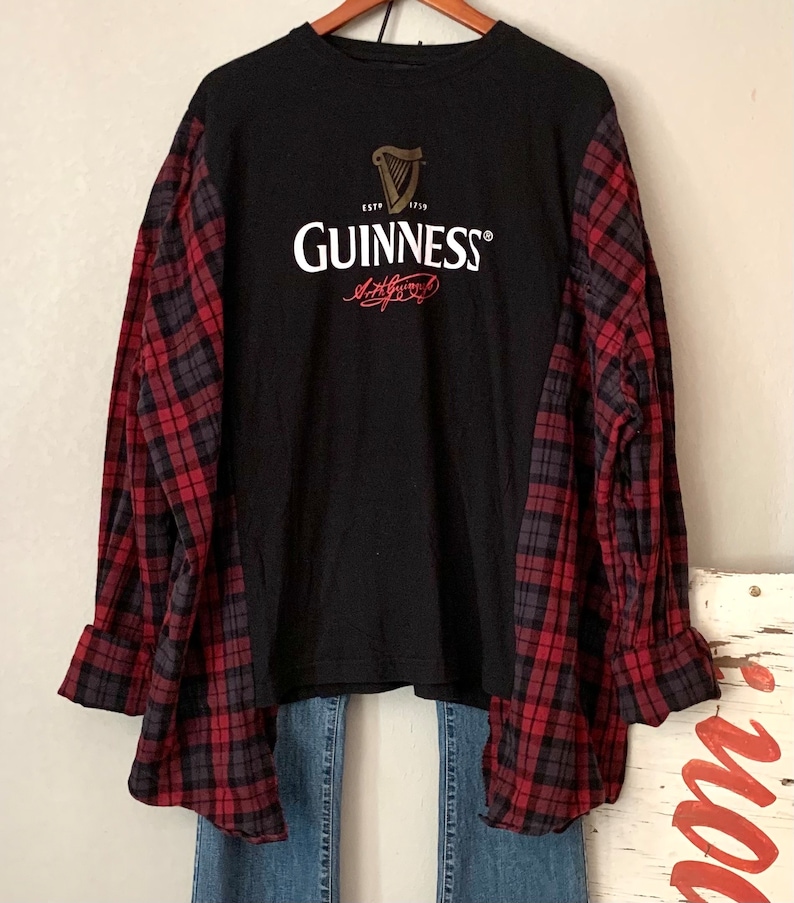 May include: A black t-shirt with a red and black checked shirt layered over it. The t-shirt features a Guinness logo with the text "ESTD 1778 GUINNESS" and a signature.