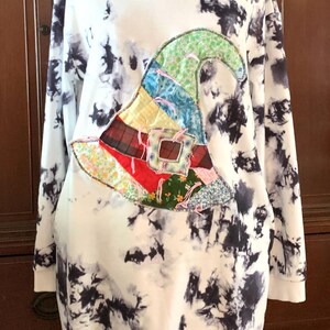 May include: A white hooded sweatshirt with a black tie-dye pattern. A colorful patchwork witch hat is appliqued on the front of the sweatshirt.