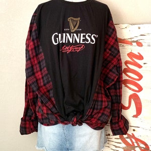 May include: Black long-sleeved shirt with a red and black checked flannel tied around the waist. The shirt features a white Guinness logo with a gold harp and the text "ESTD 1759 GUINNESS Arthur Guinness".