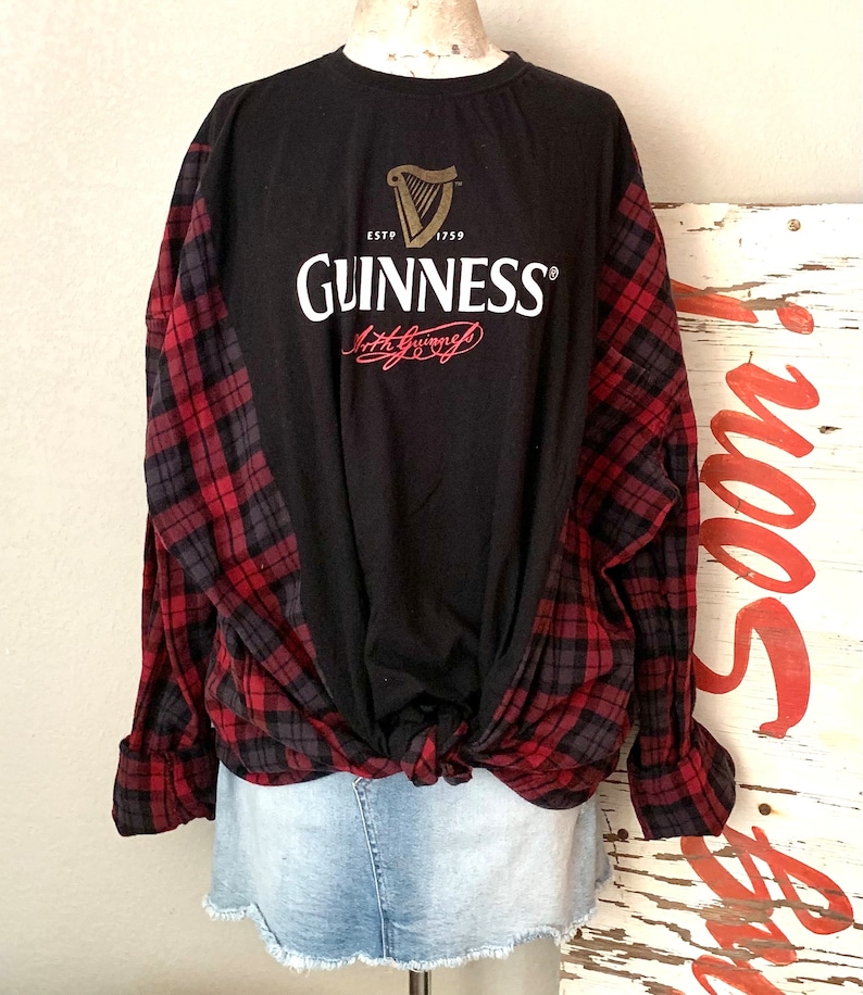 May include: Black long-sleeved shirt with a red and black checked flannel tied around the waist. The shirt features a white Guinness logo with a gold harp and the text "ESTD 1759 GUINNESS Arthur Guinness".