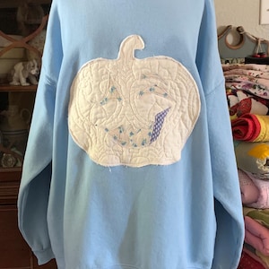May include: A light blue sweatshirt with a white quilted pumpkin appliqué on the front. The pumpkin has a stitched-on face with a wide smile and two eyes.