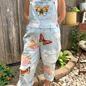 May include: Light blue denim overalls with butterfly and floral patches. The overalls have distressed details, rolled cuffs, and colorful button accents. Patches include butterflies and floral designs.