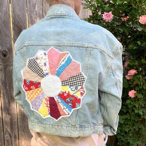 May include: A light blue denim jacket with a large, colorful patchwork flower design on the back. The flower is made of various fabrics in shades of red, pink, yellow, blue, and green.