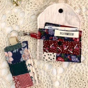 May include: A patchwork fabric card holder and matching keychain. The card holder is open, revealing cards and a label that reads "BLUEMOON." The keychain has a silver clasp and a fabric loop. The fabrics feature floral, star, and geometric patterns in red, blue, and green.