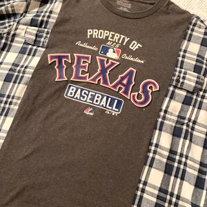 Puede incluir: Camiseta gris con un gr&aacute;fico "Property of MLB Collection Texas Baseball" en azul, rojo y blanco. La camiseta est&aacute; parcialmente cubierta por una camisa de franela a cuadros azules y blancos.