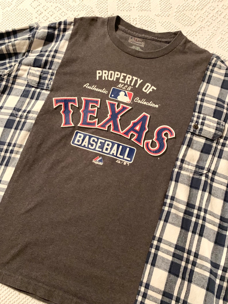 Puede incluir: Camiseta gris con un gr&aacute;fico "Property of MLB Collection Texas Baseball" en azul, rojo y blanco. La camiseta est&aacute; parcialmente cubierta por una camisa de franela a cuadros azules y blancos.