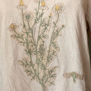 Puede incluir: Una camiseta blanca con una aplicaci&oacute;n floral verde y amarilla. La aplicaci&oacute;n es un grupo de flores con tallos y hojas verdes.