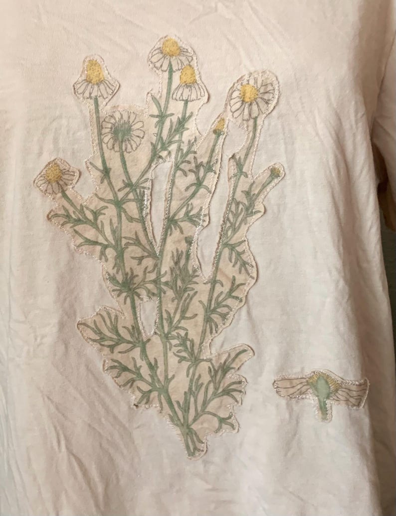 Puede incluir: Una camiseta blanca con una aplicaci&oacute;n floral verde y amarilla. La aplicaci&oacute;n es un grupo de flores con tallos y hojas verdes.