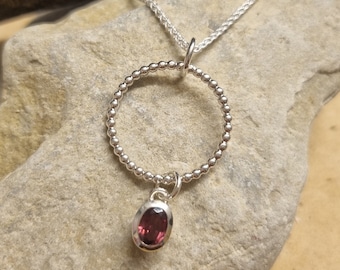 May include: A silver necklace with a circle pendant and a small oval-shaped red gemstone hanging from the bottom.