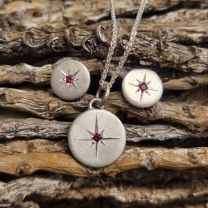 Puede incluir: Un conjunto de collar y pendientes de plata con un diseño de estrella y una piedra roja en el centro de cada pieza.