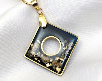 May include: A gold chain necklace with a square pendant. The pendant is made of dark blue resin with gold flecks and a circular cutout in the center.
