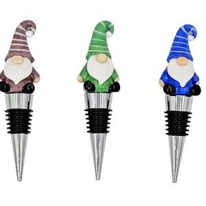 May include: Three gnome-shaped bottle stoppers. The gnome on the left is purple with white stripes, the middle gnome is green with white stripes, and the gnome on the right is blue with white stripes. All three gnomes have white beards and black hats.