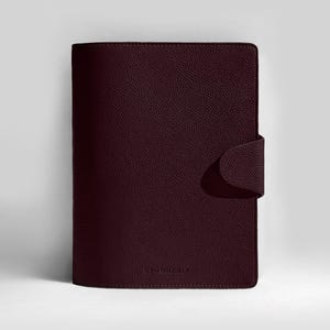 May include: A burgundy leather journal with a textured surface. The journal has a rounded flap closure on the right side. The journal is closed and upright against a white background.